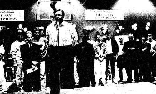 A man speaks at a microphone in a gym full of people
