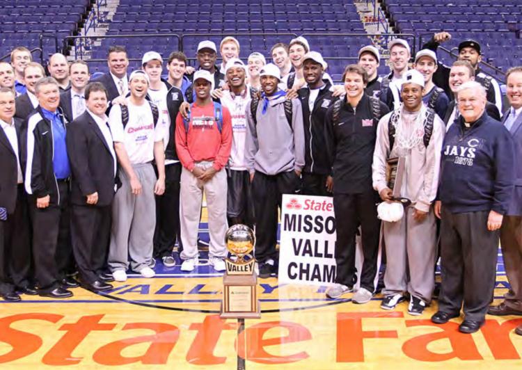 A basketball team stands together on the court in front of a trophy