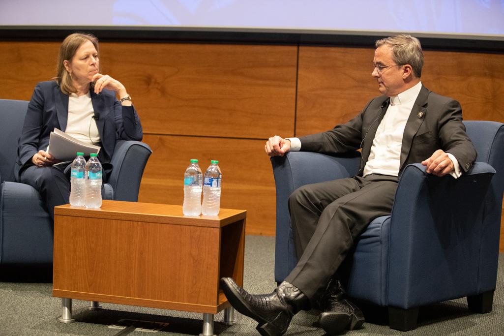 Two people sit in chairs on a stage and talk