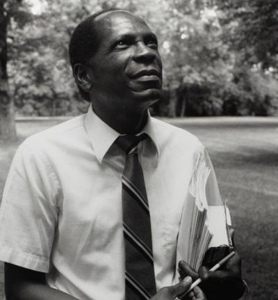 A man, in a shirt and tie, looks up as he carries notebooks under his arm