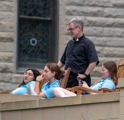 A Jesuit stands next to students sitting on rocking chairs