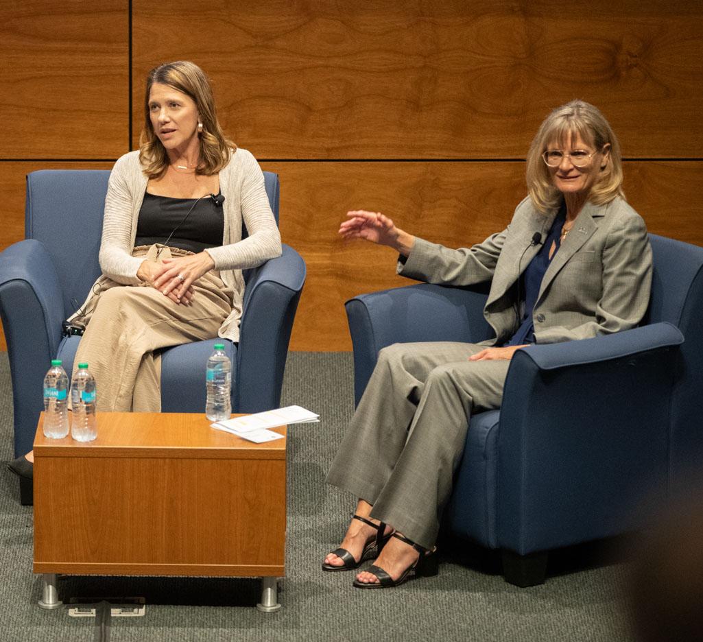 Two people sit in chairs on a stage and talk