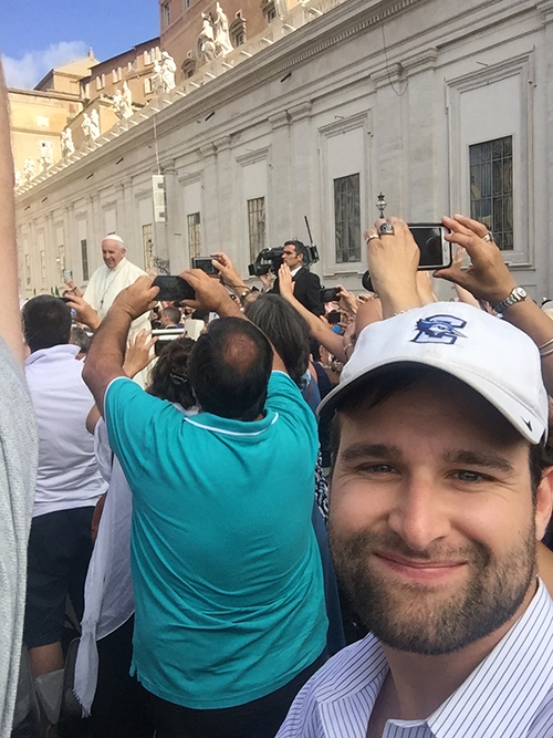 Zach Perry snapping a selfie with Pope Francis in the background at St. Peter's Square in Vatican City.