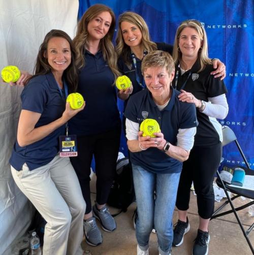 Five women pose together for a photo, with four of the women holding yellow softballs