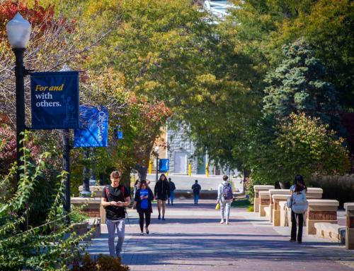 Students walking on campus with a "For and with others" sign