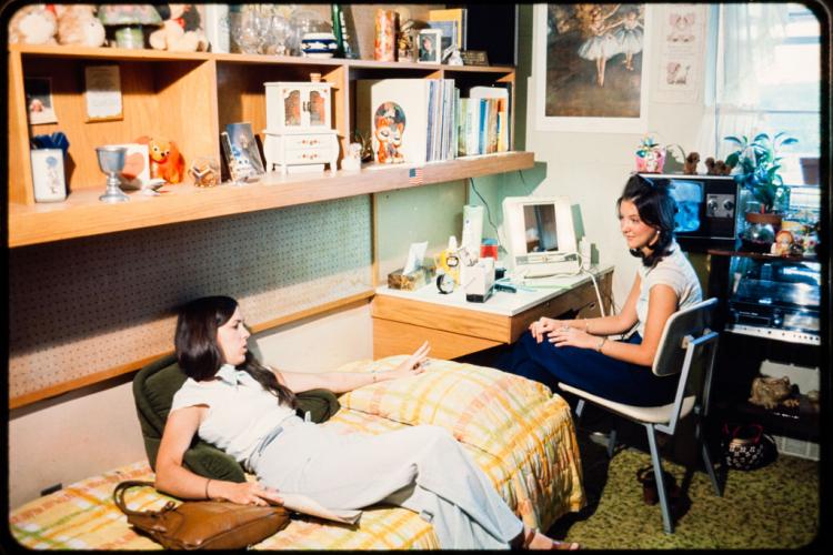 A woman sits on a chair talking a friend lounging on her bed.