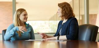 A student and faculty member talk while sitting at a table.